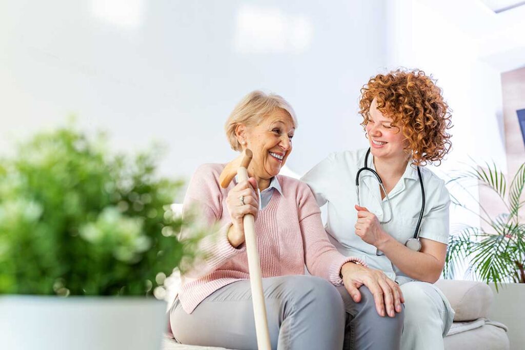 Older adult reading in a sunny private room while a caregiver respectfully assists nearby.